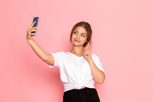 Una vista frontal joven y bella mujer en camisa blanca posando con expresión divertida y tomando una selfie