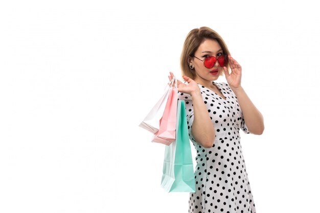 Una vista frontal joven y bella mujer en vestido de lunares en blanco y negro con gafas de sol rojas con paquetes de compras posando