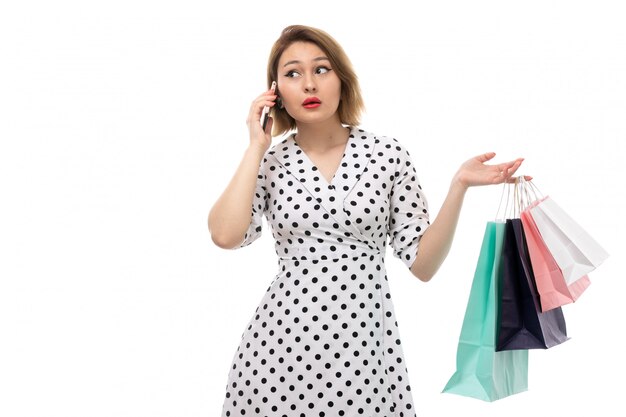 Una vista frontal joven y bella mujer en vestido de lunares en blanco y negro con paquetes de compras hablando por teléfono