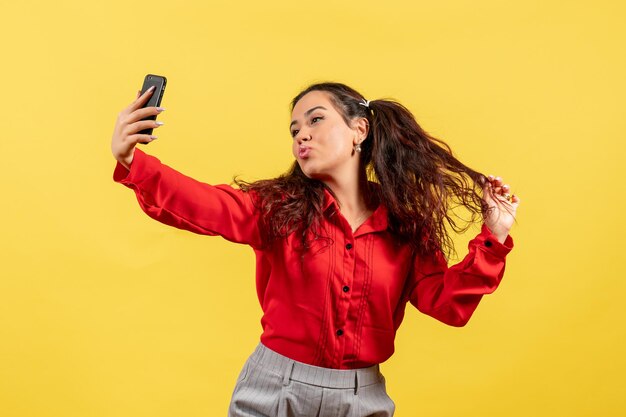 Vista frontal joven en blusa roja con cabello lindo tomando selfie en escritorio amarillo niño niña juventud inocencia color niño