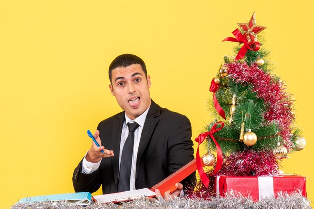 Vista frontal joven con la boca abierta sentado en la mesa mostrando documentos árbol de Navidad y regalos