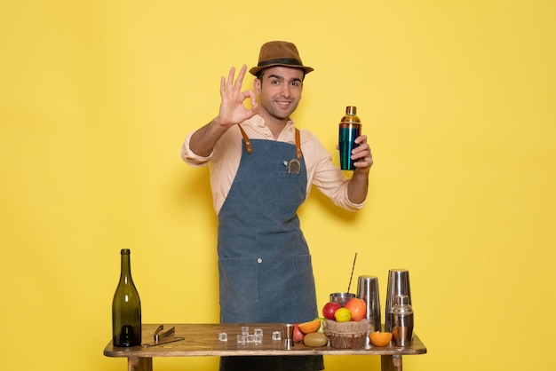 Vista frontal joven camarero frente a la mesa con bebidas sosteniendo agitador sobre fondo amarillo