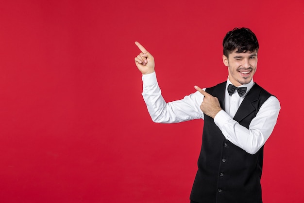 Foto gratuita vista frontal del joven camarero masculino divertido en uniforme con pajarita en el cuello y apuntando hacia arriba en el lado derecho de la pared roja