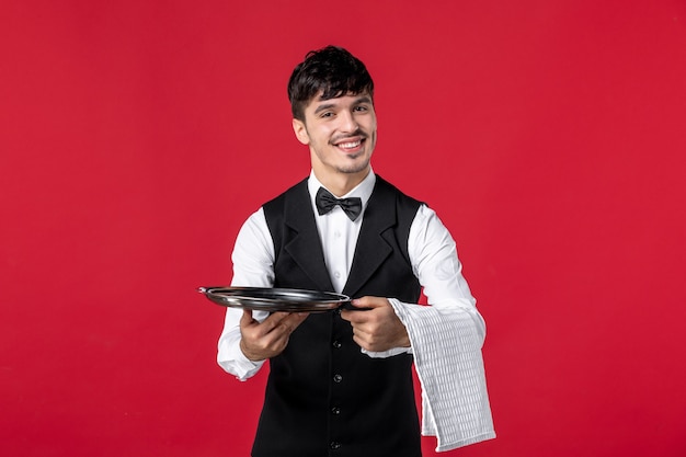 Foto gratuita vista frontal del joven camarero masculino sonriente en uniforme con pajarita en el cuello sosteniendo la bandeja y la toalla en la pared roja