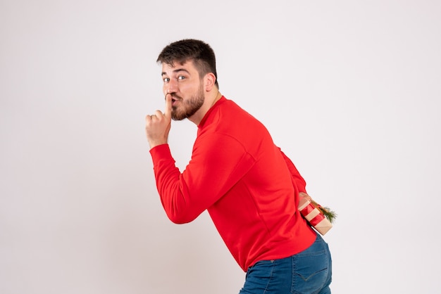 Foto gratuita vista frontal del joven con camisa roja escondiendo el regalo de navidad detrás de su espalda en la pared blanca