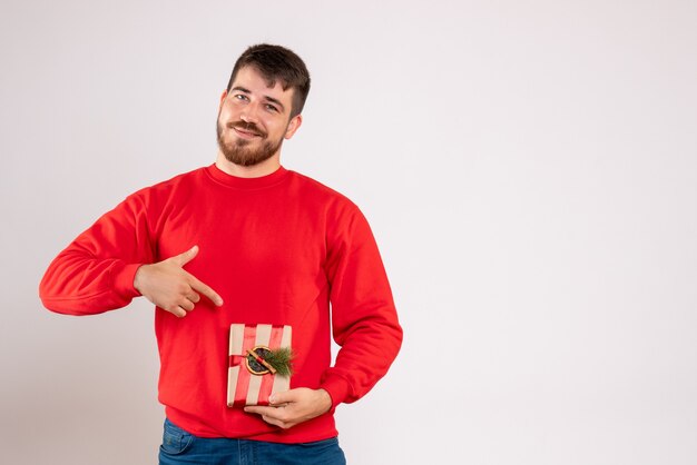 Vista frontal del joven en camisa roja con pequeño regalo de Navidad en la pared blanca