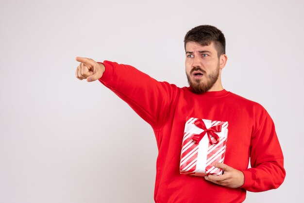 Vista frontal del joven en camisa roja con regalo de Navidad apuntando en la pared blanca