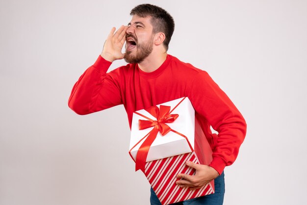 Vista frontal del joven en camisa roja con regalo de Navidad en caja y gritando en la pared blanca