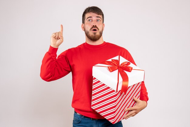 Vista frontal del joven en camisa roja con regalo de Navidad en caja en pared blanca