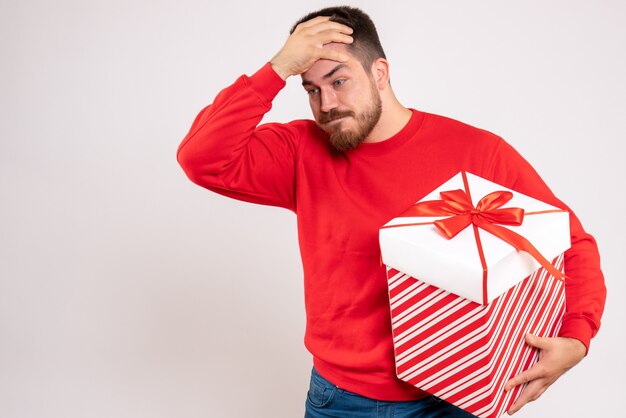Vista frontal del joven en camisa roja con regalo de Navidad en caja en pared blanca
