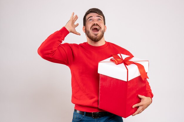 Vista frontal del joven en camisa roja con regalo de Navidad en caja en pared blanca