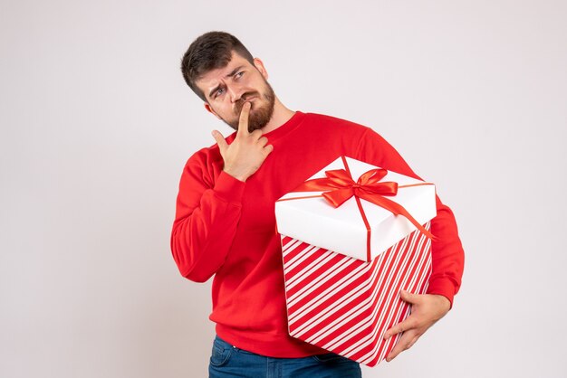 Vista frontal del joven en camisa roja con regalo de Navidad en caja pensando en pared blanca