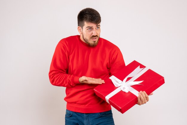 Vista frontal del joven en camisa roja con regalo de Navidad en la pared blanca