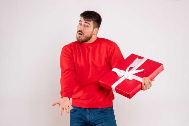 Vista frontal del joven en camisa roja con regalo de Navidad en la pared blanca