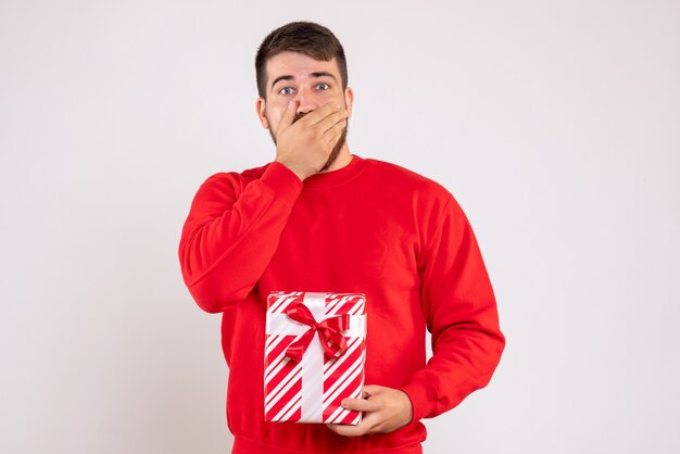 Vista frontal del joven en camisa roja con regalo de Navidad en la pared blanca