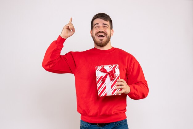 Vista frontal del joven en camisa roja con regalo de Navidad en la pared blanca