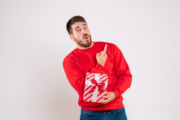 Vista frontal del joven en camisa roja con regalo de Navidad en la pared blanca