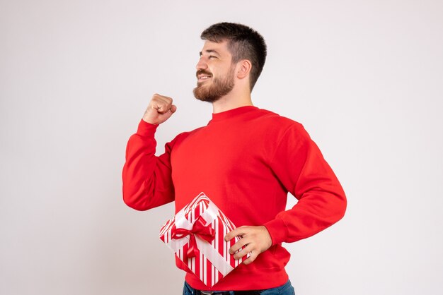 Vista frontal del joven en camisa roja con regalo de Navidad en la pared blanca