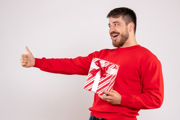 Vista frontal del joven en camisa roja con regalo de Navidad en la pared blanca