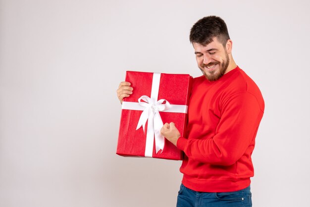 Vista frontal del joven en camisa roja con regalo de Navidad sonriendo en la pared blanca