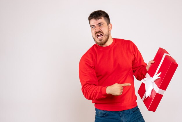 Vista frontal del joven con camisa roja sosteniendo el regalo de Navidad y discutiendo con alguien en la pared blanca