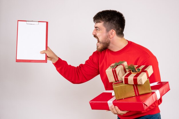 Vista frontal del joven en camisa roja sosteniendo regalos de Navidad discutiendo sobre pared blanca