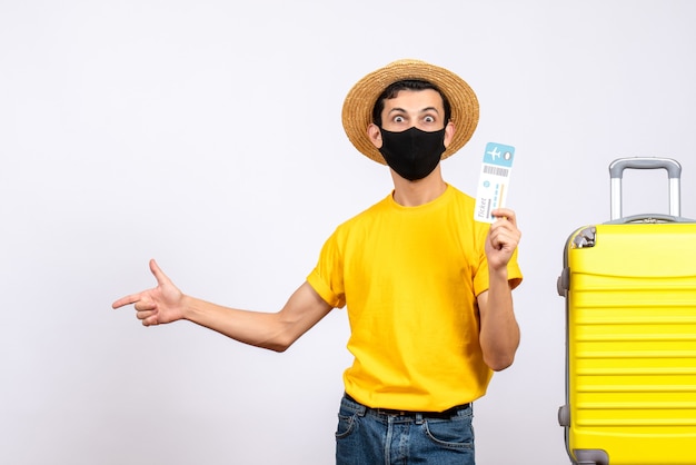 Vista frontal joven en camiseta amarilla de pie cerca de la maleta amarilla sosteniendo el boleto de viaje