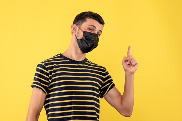 Vista frontal joven en camiseta a rayas blanco y negro apuntando con el dedo hacia arriba sobre fondo amarillo aislado