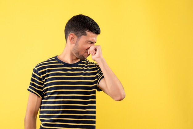 Vista frontal joven en camiseta a rayas blanco y negro cerrando la nariz sobre fondo amarillo aislado