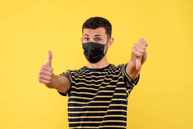 Vista frontal joven en camiseta a rayas blanco y negro haciendo pulgar hacia arriba y hacia abajo firmar sobre fondo amarillo