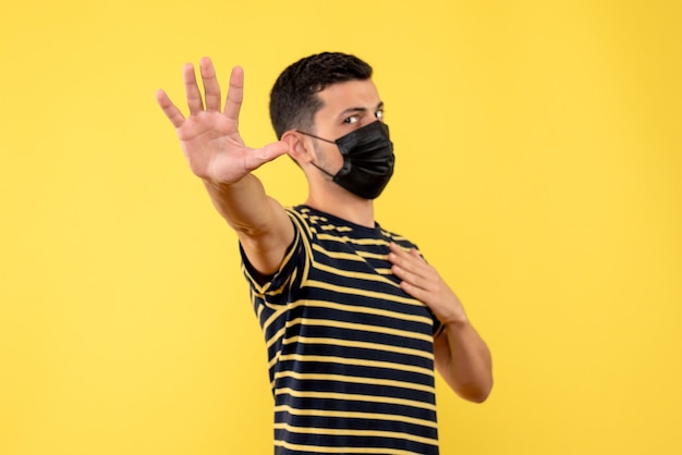 Vista frontal joven en camiseta a rayas blanco y negro haciendo señal de stop sobre fondo amarillo