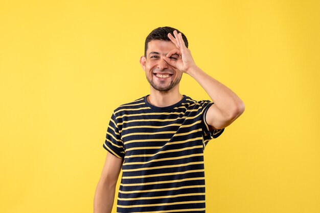 Vista frontal joven en camiseta a rayas blanco y negro poniendo signo okey delante de sus ojos sobre fondo amarillo aislado