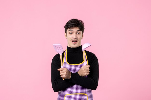 Vista frontal joven en capa con cucharas sobre fondo rosa color comida cocina uniforme trabajo masa cocina profesión