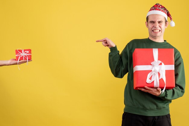 Vista frontal joven cerrando los ojos apuntando al regalo en la mano femenina en amarillo