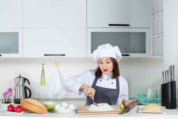Vista frontal de la joven chef femenina concentrada en uniforme preparando la comida en la cocina blanca