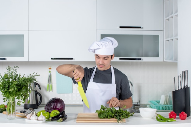 Vista frontal joven chef con gorro de cocinero y delantal cortando verduras