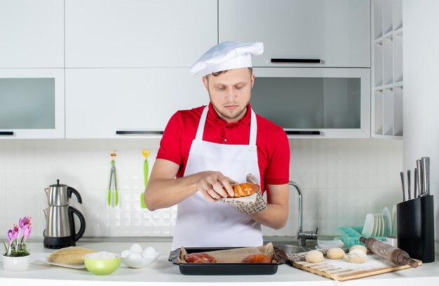 Vista frontal del joven chef masculino ocupado vistiendo titular sosteniendo uno de los pasteles recién horneados en la cocina blanca