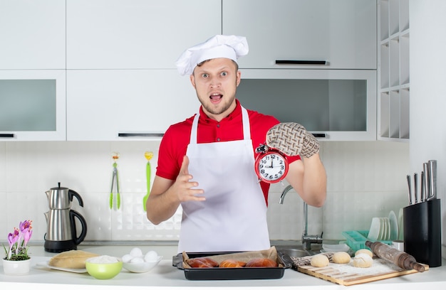 Vista frontal del joven chef masculino sorprendido con soporte sosteniendo el reloj y haciendo el gesto de ok en la cocina blanca