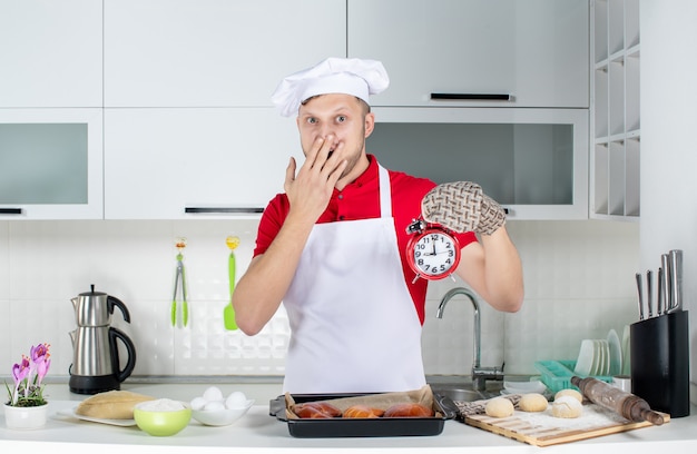 Vista frontal del joven chef masculino sorprendido vistiendo titular sosteniendo el reloj en la cocina blanca