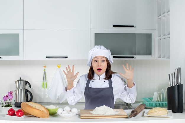 Vista frontal de la joven chef mujer asustada en uniforme de pie detrás de la mesa con alimentos de tabla de cortar en la cocina blanca