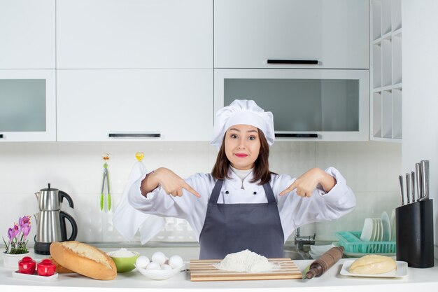 Vista frontal de la joven chef mujer sonriente apuntando con su uniforme en la cocina blanca