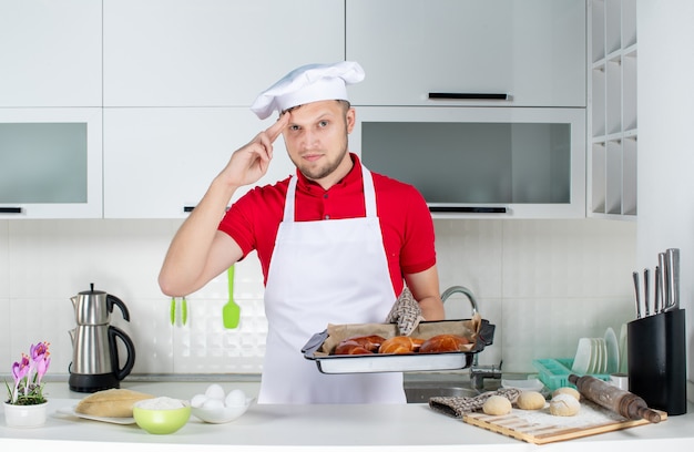 Vista frontal del joven chef con soporte sosteniendo pasteles recién horneados y sintiéndose orgulloso en la cocina blanca