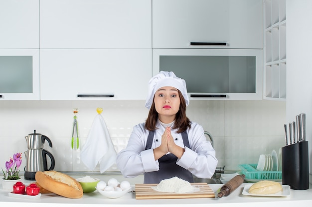 Vista frontal de la joven chef en uniforme rezando por algo en la cocina blanca