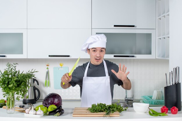 Vista frontal joven chef en uniforme sosteniendo verduras de corte de cuchillo