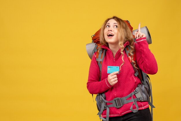 Vista frontal de la joven chica viajera sonriente en máscara médica recogiendo su equipaje con tarjeta bancaria apuntando hacia arriba