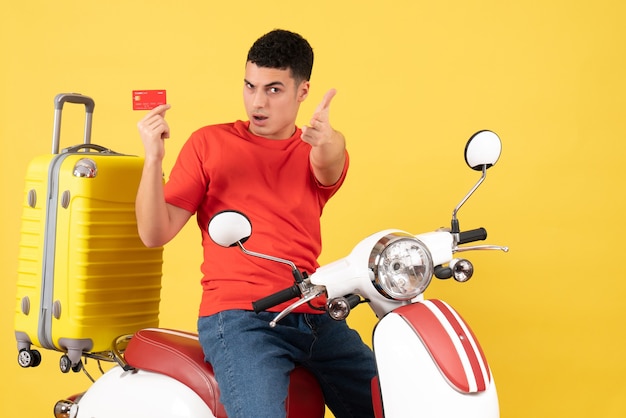 Vista frontal joven en ciclomotor con tarjeta de crédito señalando gesto de pistola con el dedo a la cámara