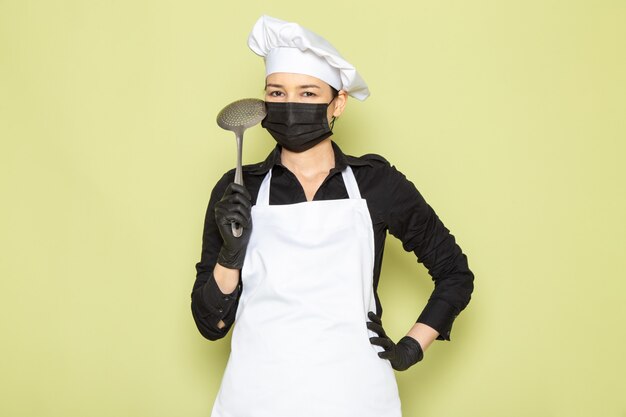 Una vista frontal joven cocinera en camisa negra capa de cocinero blanco gorra blanca en guantes negros máscara negra posando sosteniendo una cuchara de plata grande