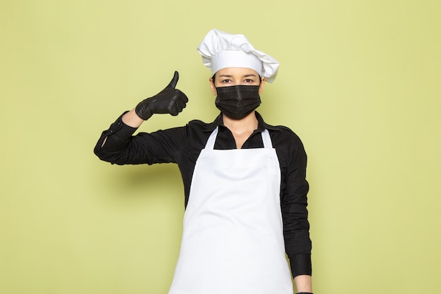 Foto gratuita una vista frontal joven cocinera en camisa negra capa de cocinero blanco gorra blanca posando en guantes negros máscara negra posando