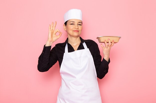 Una vista frontal joven cocinera en camisa negra capa de cocinero blanco gorra blanca posando sosteniendo tazón de leche marrón