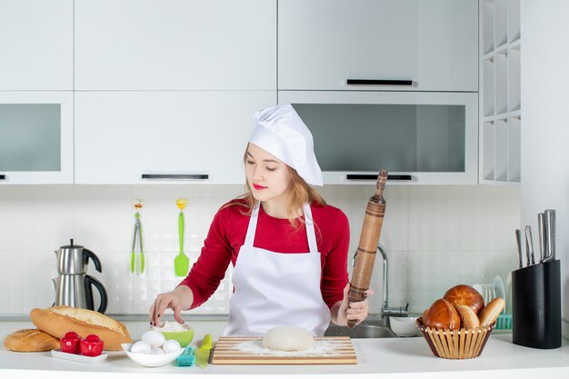 Vista frontal joven cocinera espolvorear harina a la masa en la tabla de cortar sosteniendo el rodillo en la cocina